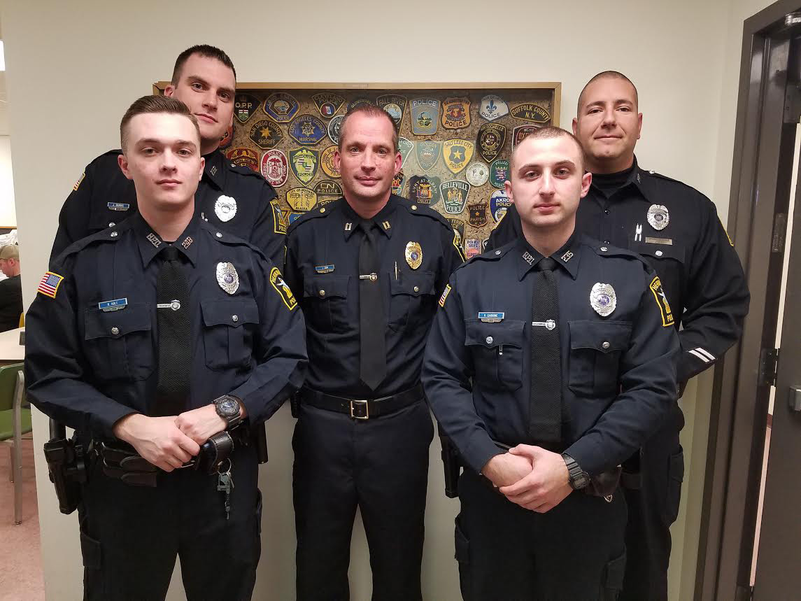Five police officers pose in front of a bulletin board