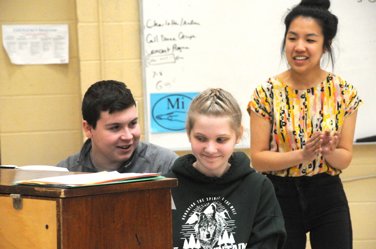 Two students sit at a piano while a woman looks on