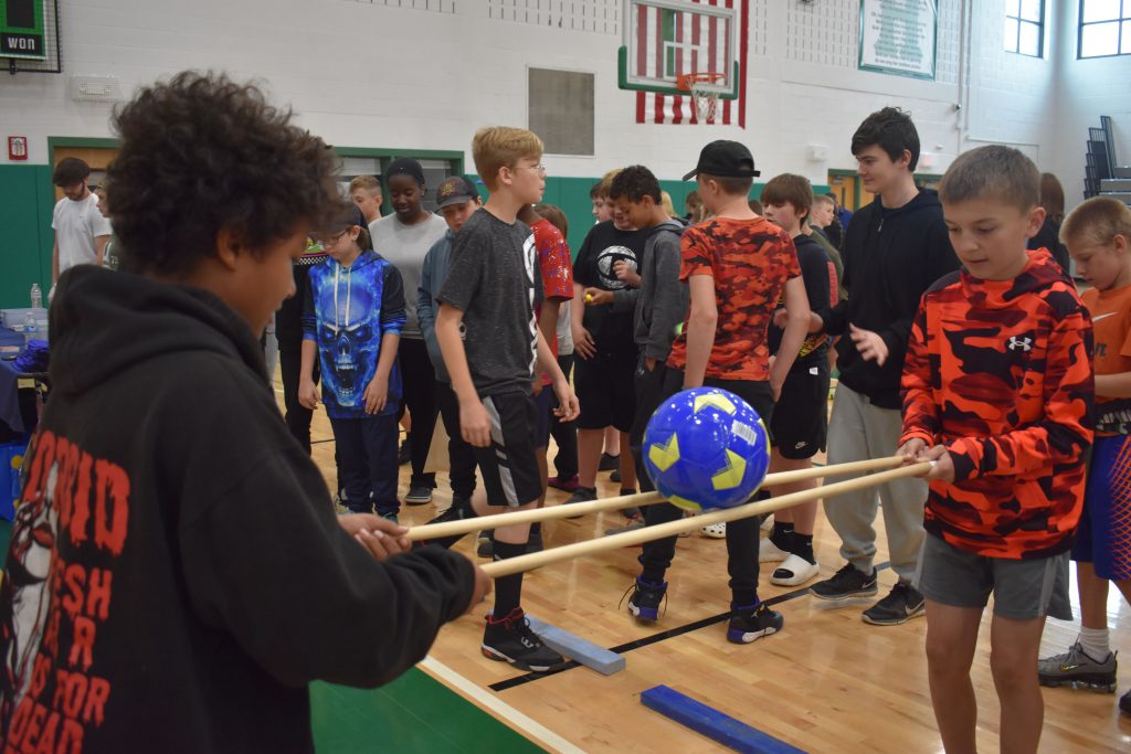 Two students holding stick and balancing a ball in the gym as other students look on