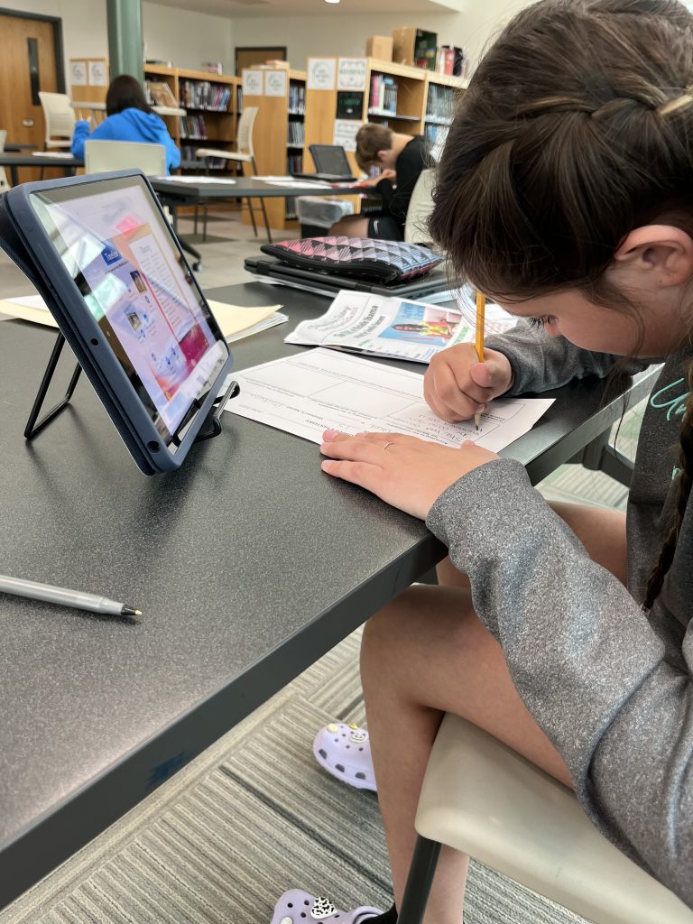 Seventh grade officer writing while looking at a newspaper from a project about the magic of women's history