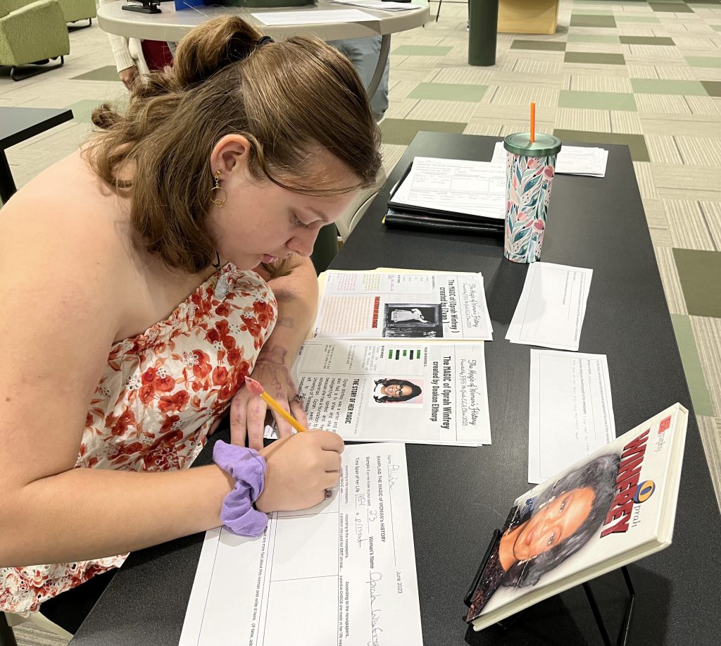 Seventh grade officer writing while looking at a newspaper from a project about the magic of women's history