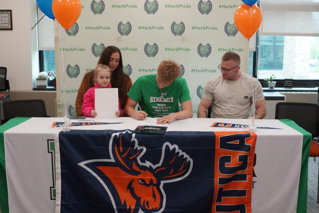 Kaleb Davis and three others at his Utica University signing with a Utica University banner on the table