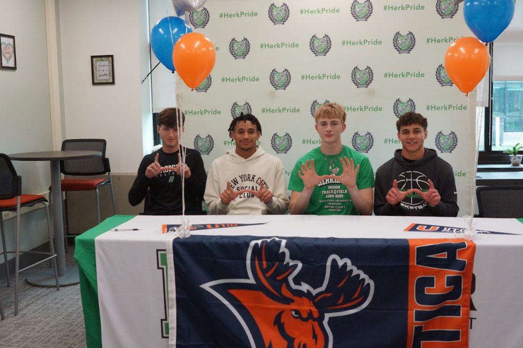 Kaleb Davis and three others at his Utica University signing with a Utica University banner on the table and they're making U shapes with their hands