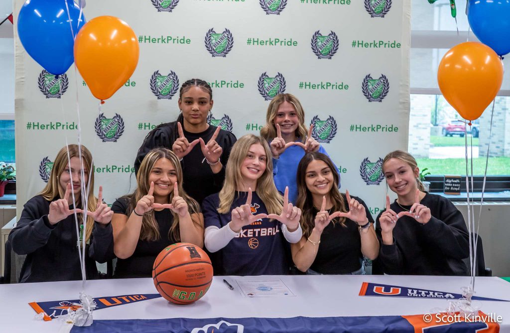Madison Marusic and six others all making a U sign with their hands at her Utica University signing with a Utica University banner on the table