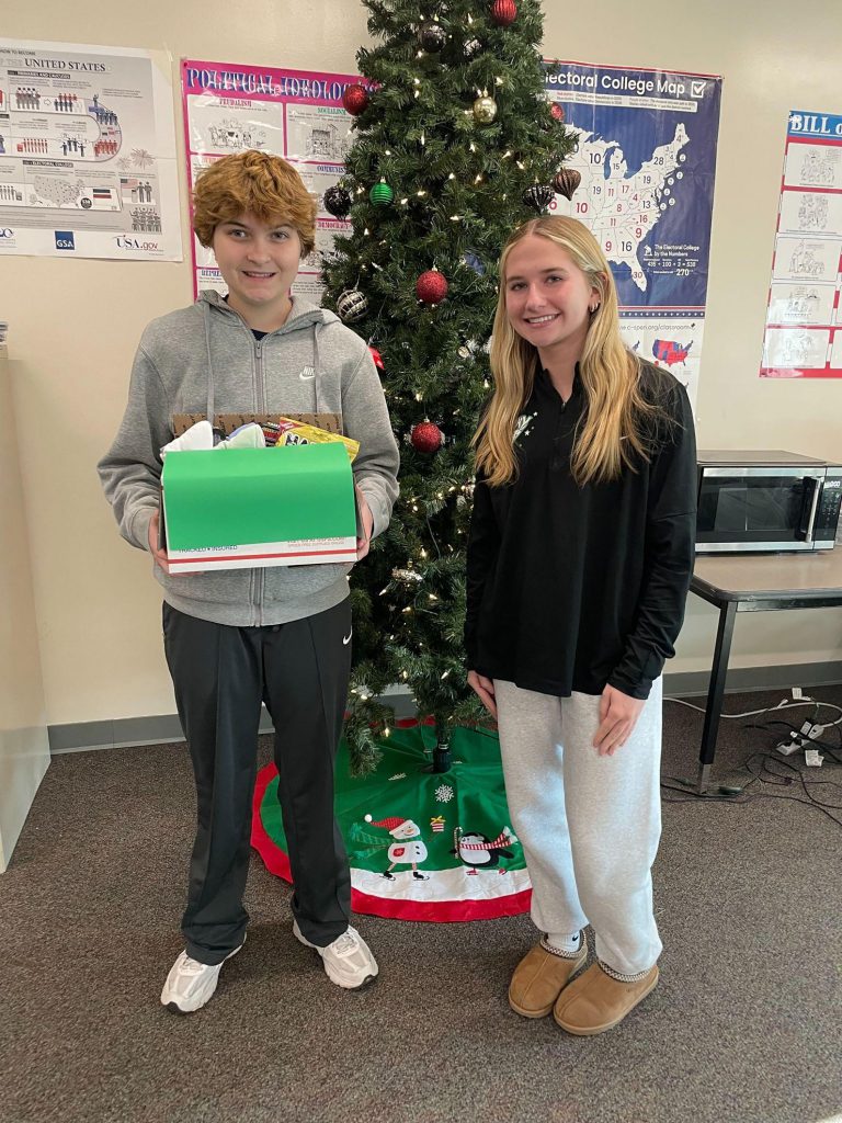 Two students posing by Christmas tree with donations for Holiday Project