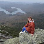 Emily Schumske sitting on large rocks above a a wide landscape with water and more
