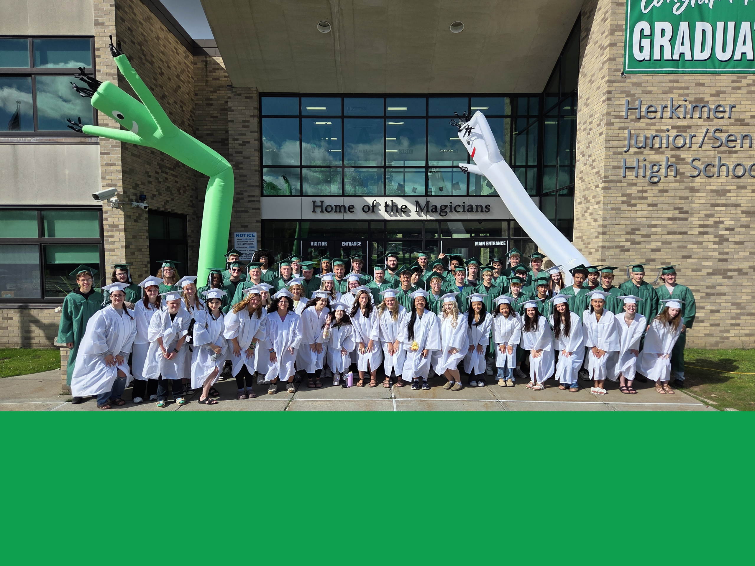 Class of 2026 seniors in caps and gowns in front of the Middle-High School with large balloon people