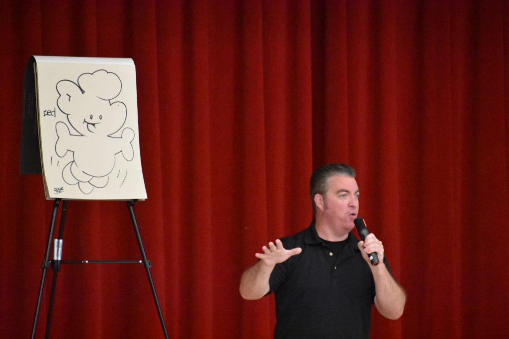 Cartoonist Duane Abel speaking on stage in front of an illustration he made