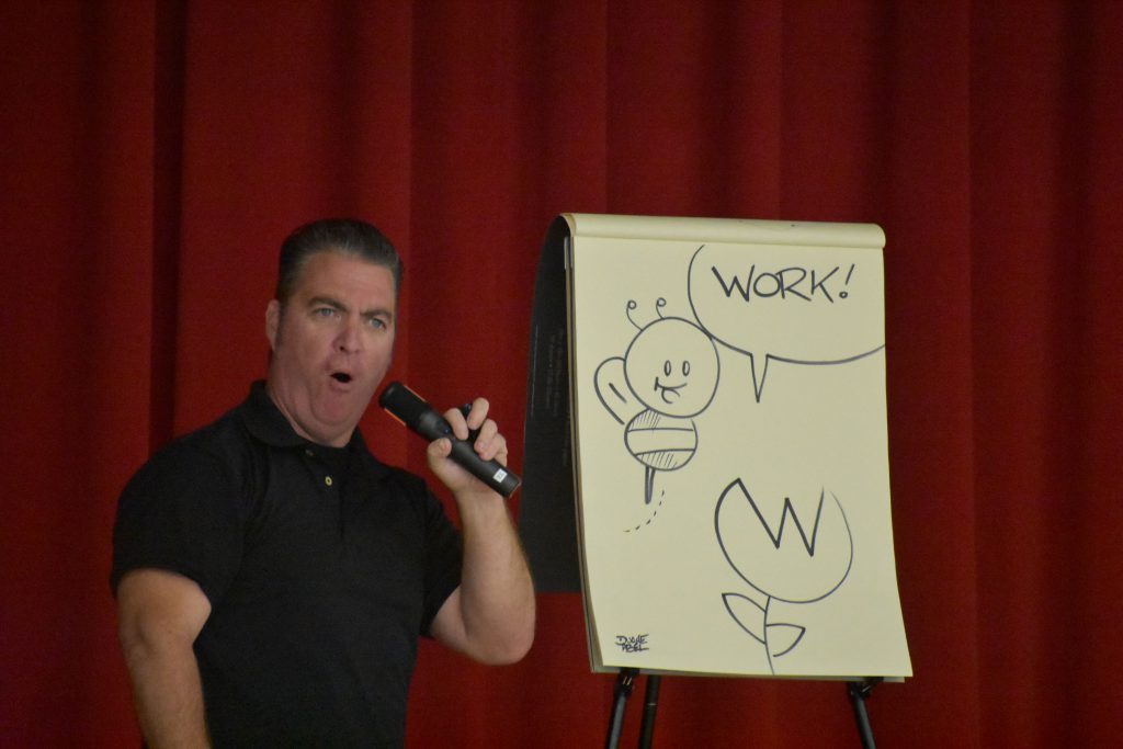 Cartoonist Duane Abel speaking on stage in front of an illustration he made