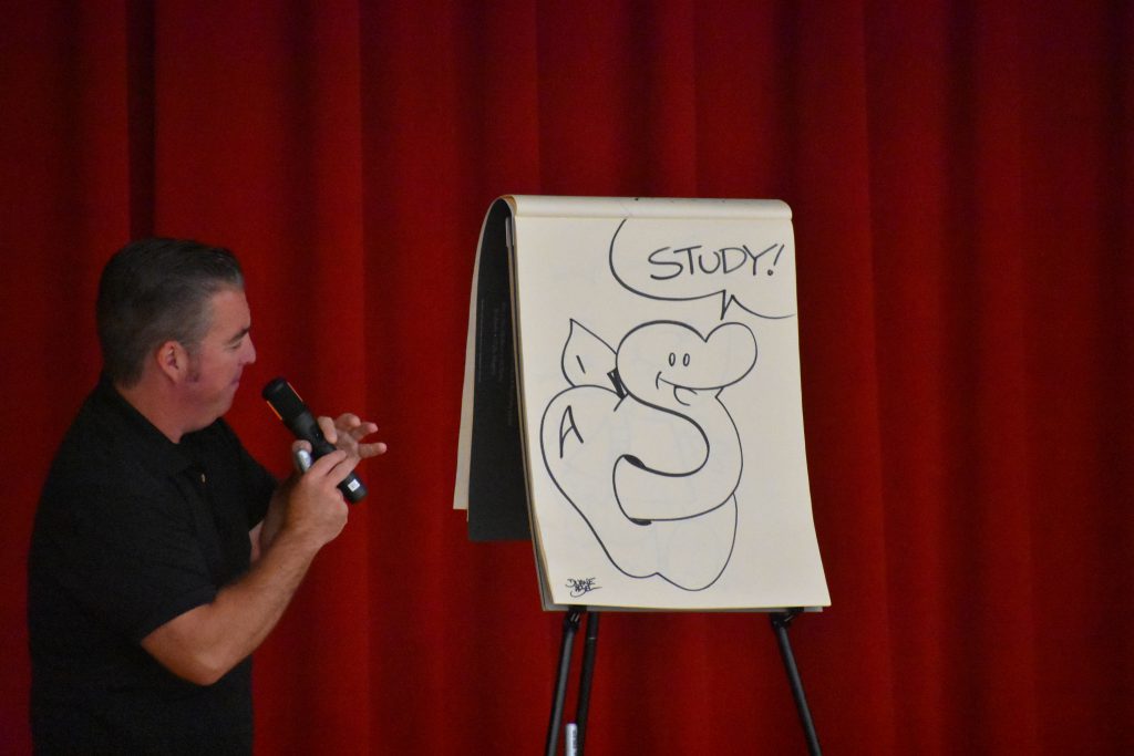 Cartoonist Duane Abel speaking on stage in front of an illustration he made