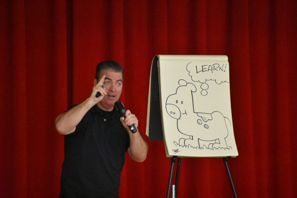 Cartoonist Duane Abel speaking on stage in front of an illustration he made