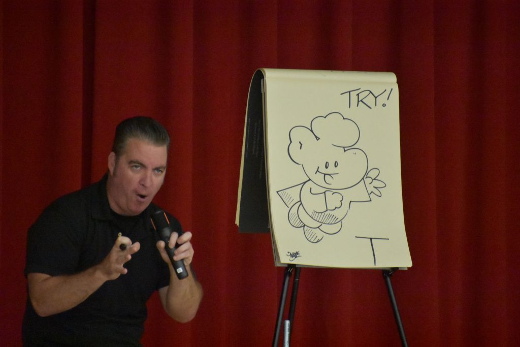 Cartoonist Duane Abel speaking on stage in front of an illustration he made
