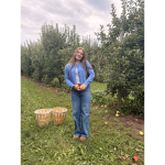 Adah Foster standing outside holding two apples with two baskets near her and apple trees behind her