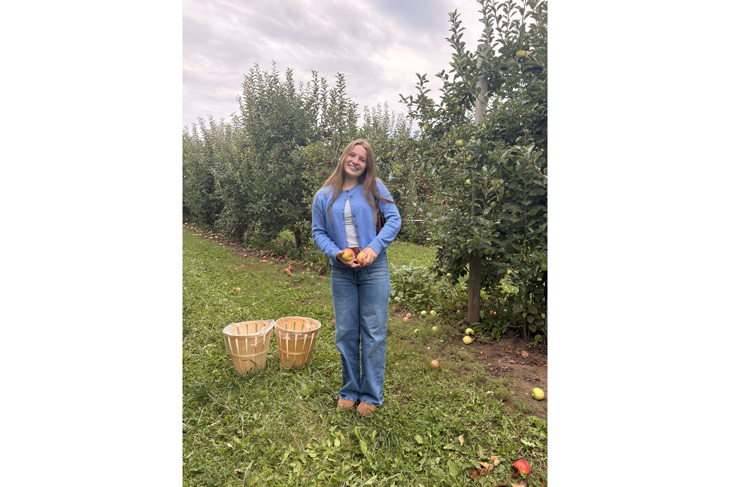 Adah Foster standing outside holding two apples with two baskets near her and apple trees behind her